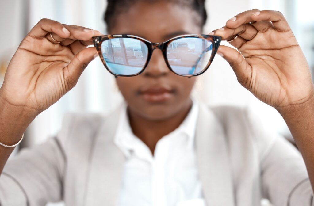 A person (not in focus) holding up a pair of glasses with blue light glare on the lenses.