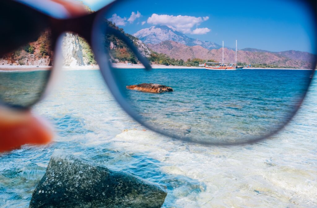 A hand holding polarized sunglasses against a blue ocean, showing enhanced color and visual clarity